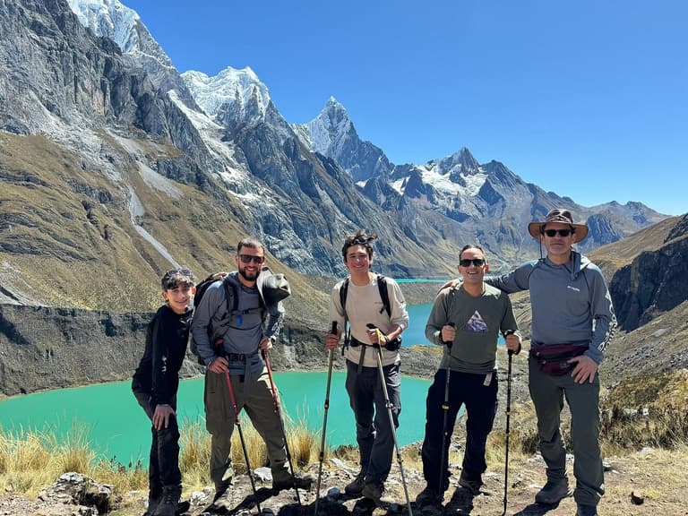 A group of five trekkers with poles and packs standing above a turquoise lake, with the snow-capped peaks of the Cordillera Huayhuash behind them
