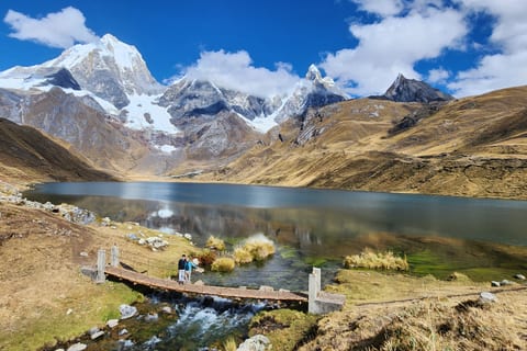 Two trekkers crossing a wooden footbridge at the outlet of a turquoise lake, snow walls rising behind, Cordillera Huayhuash