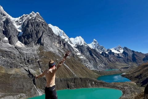 A trekker with arms raised at the Three Lakes viewpoint, three turquoise lakes below the snow walls of the Cordillera Huayhuash